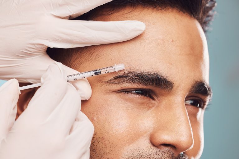 Handsome young indian man receiving a botox injection against a blue studio background. Mixed race guy getting treatment to reduce ageing and fill facial wrinkles